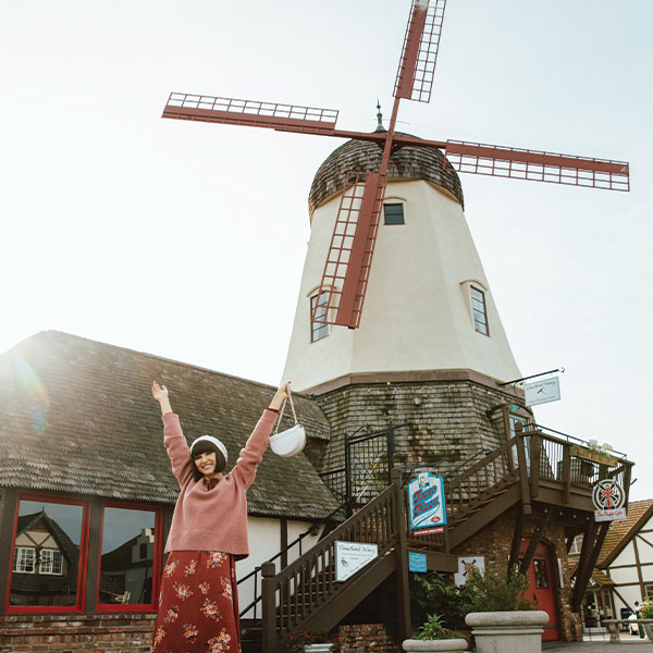 where to stay in downtown Solvang A woman in a pink sweater and red skirt stands smiling with arms raised in front of a large windmill with red blades on a sunny day, capturing a joyful moment perfect for Press & Media features.