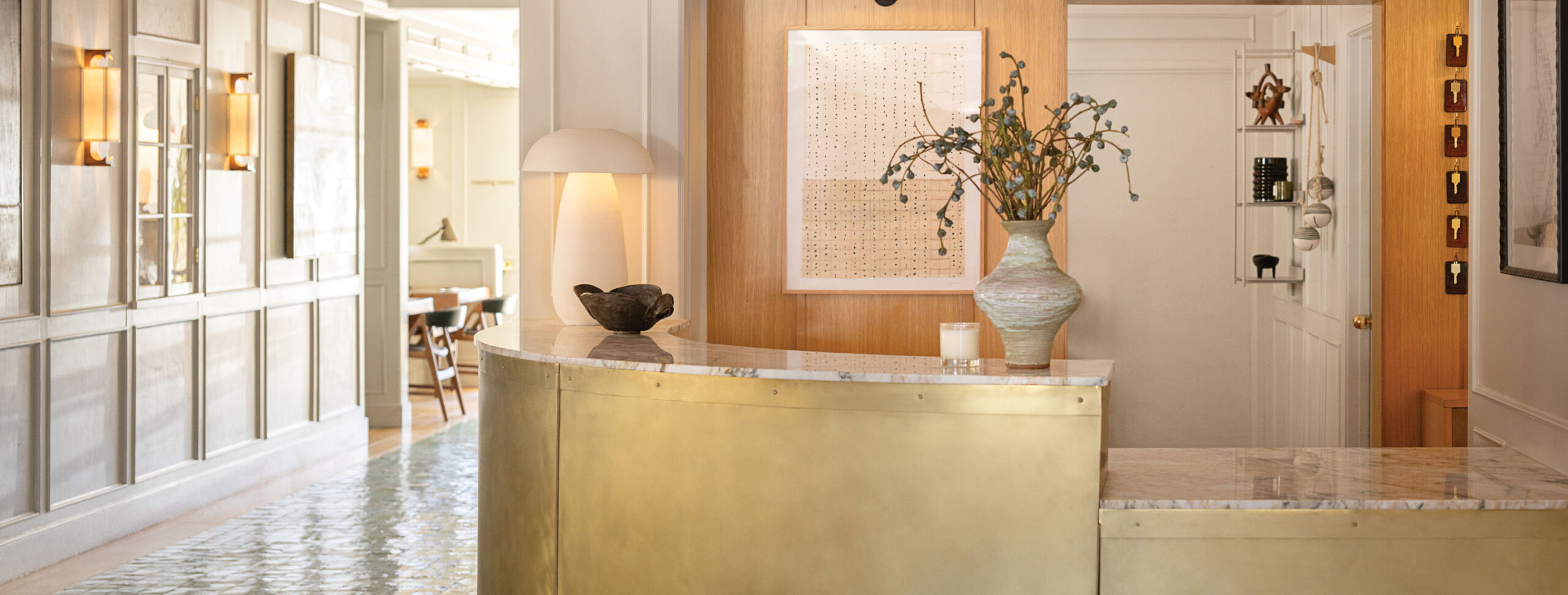 A well-lit hotel reception area features a curved brass desk with a marble top, decorative vases, modern wall art, and stylish paneling.