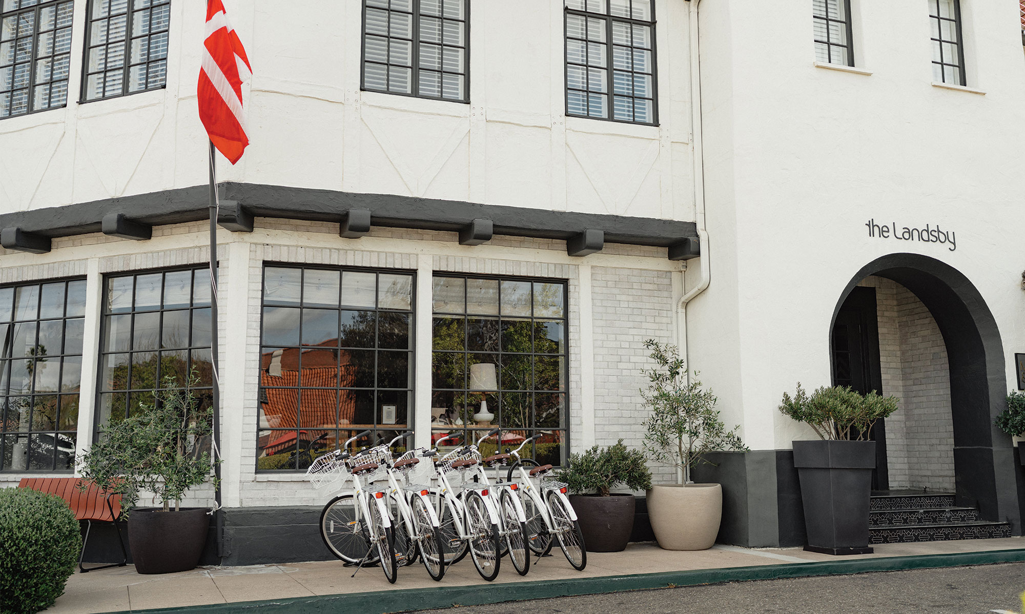 A row of white bicycles—one of the inviting hotel amenities at The Landsby—is parked outside its white building with large windows and a black awning. A red-and-white striped flag is mounted near the entrance.