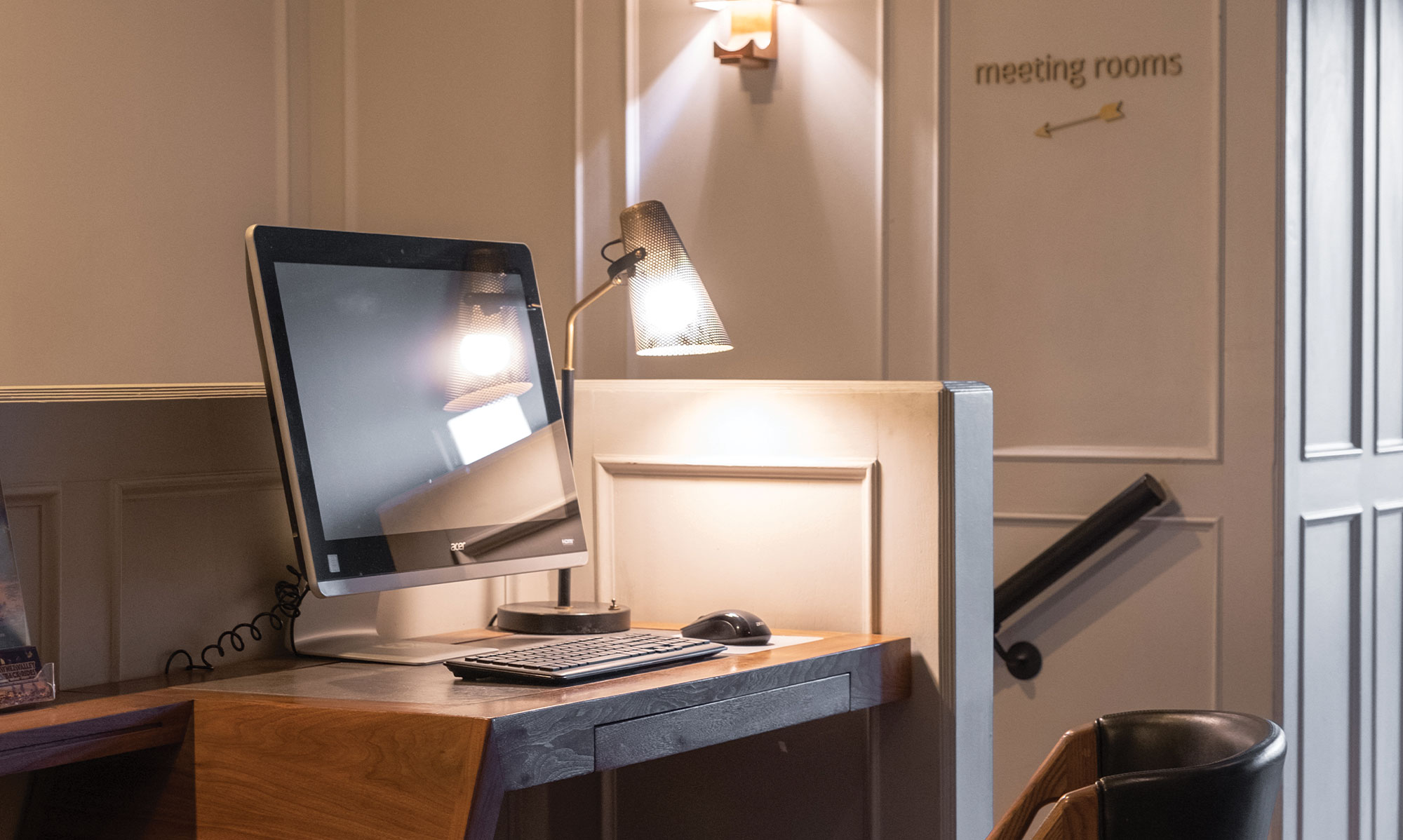 A computer with a keyboard and mouse sits on a wooden desk under a desk lamp. In the background, a sign on the wall points to meeting rooms. The setting highlights the comfortable hotel amenities at The Landsby in a quiet office or lobby area.