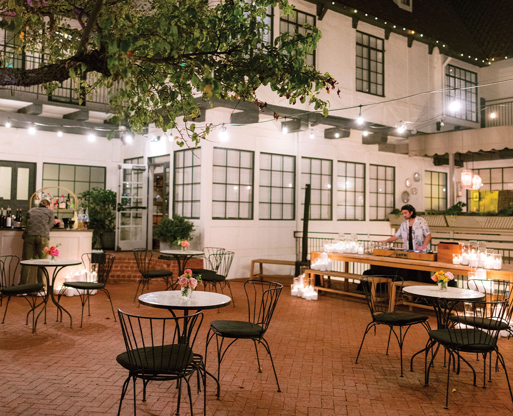 A cozy outdoor patio with round tables and black chairs, string lights overhead, and a bar setup. Perfect for gatherings, this inviting area is one of the charming event spaces in Solvang, with flowers on each table and a warm glow throughout.