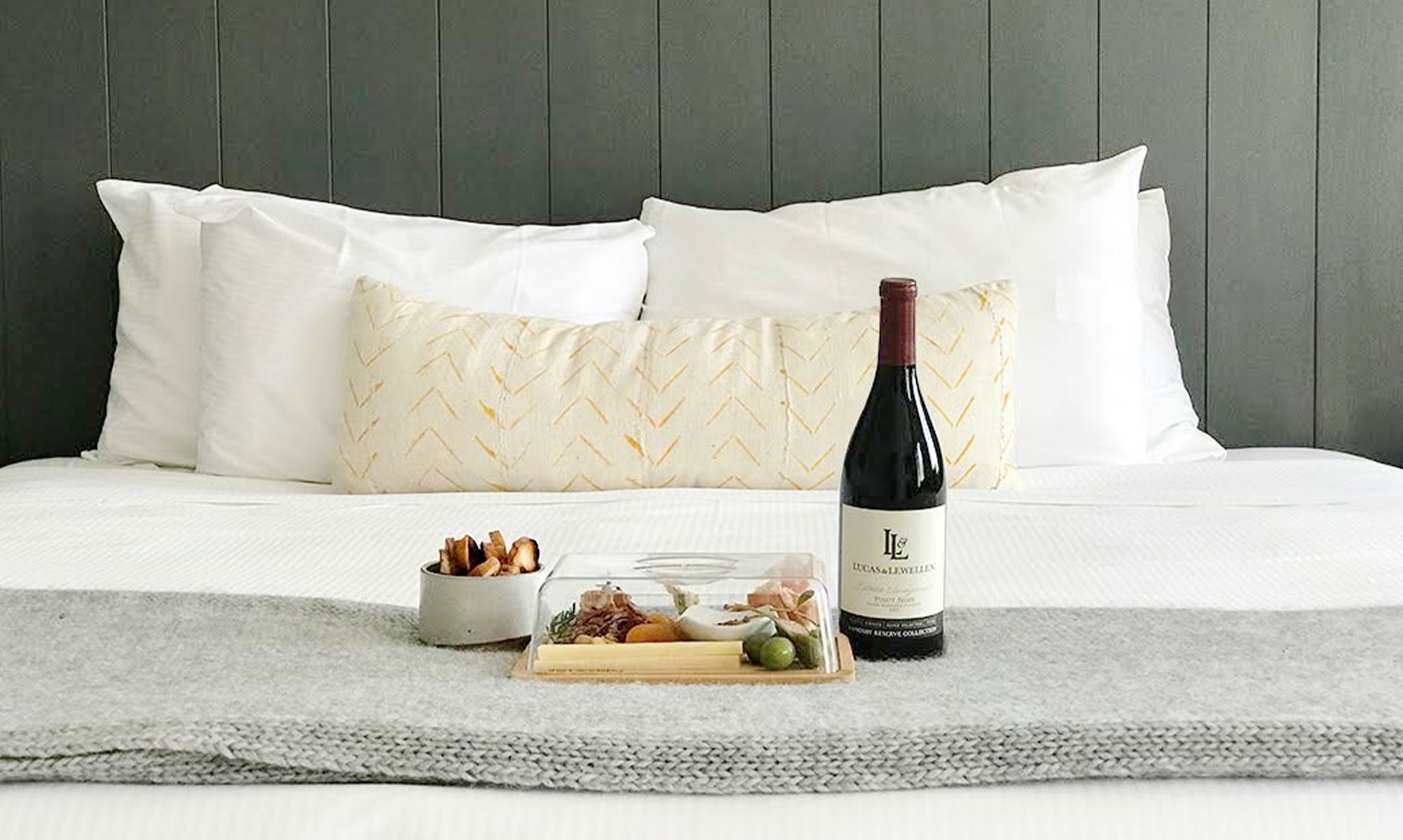 A bottle of red wine, a small bowl of snacks, and a cheese platter rest on a gray blanket atop a neatly made bed with white pillows—just some of the elegant hotel amenities at The Landsby.