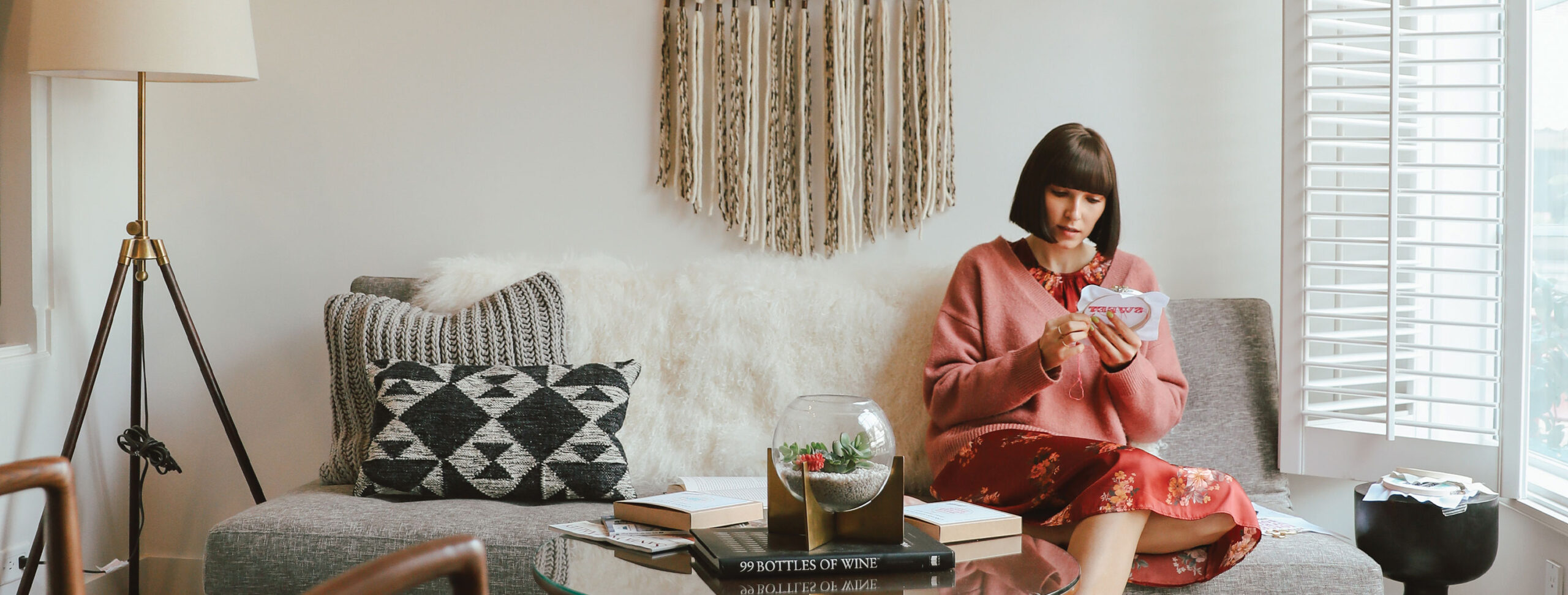 Suites at The Landsby A woman with a bob haircut sits on a gray sofa reading a card, possibly checking Frequently Asked Questions. She wears a pink sweater and red floral skirt. The room features decorative pillows, books, wall art, and large windows with white shutters.