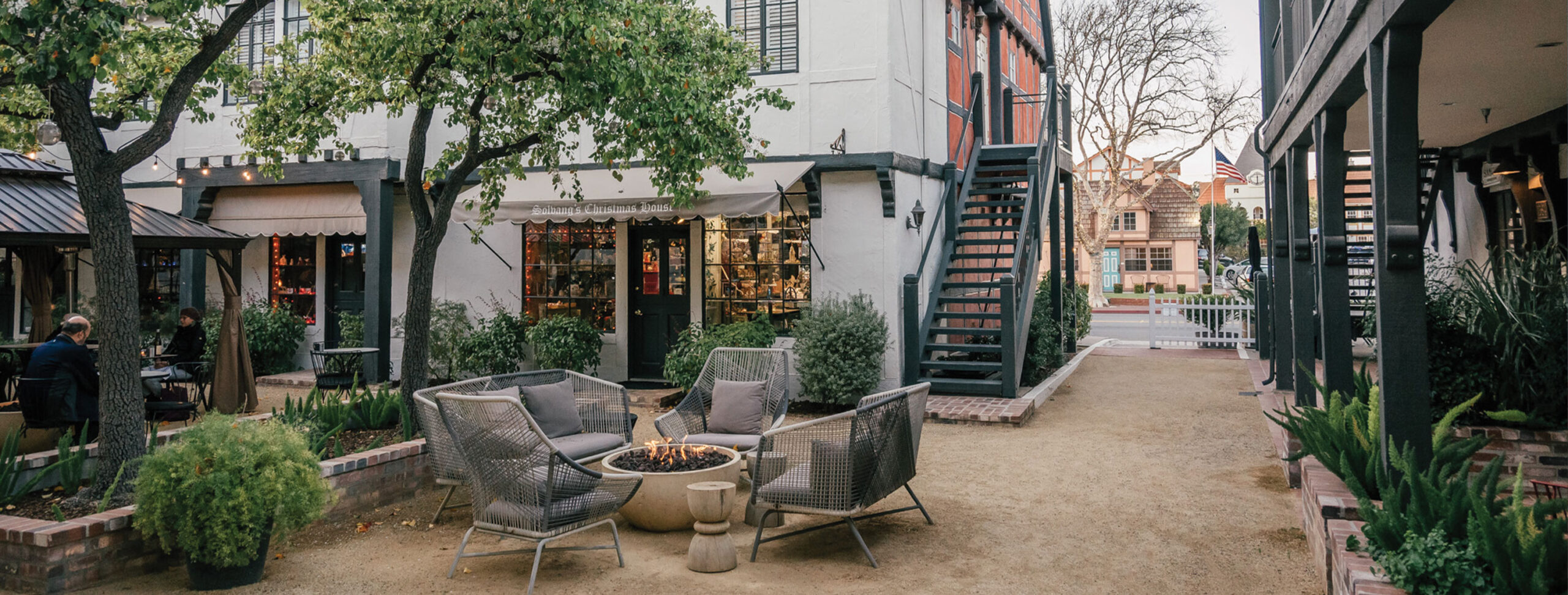Outdoor courtyard with modern chairs around a fire pit, surrounded by greenery and shops. Stairs lead to a second floor, and a path runs through the space. Trees and string lights create a cozy vibe—perfect for photos of The Landsby.
