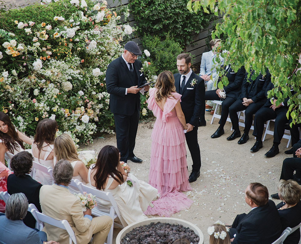 A couple stands together during an outdoor wedding ceremony at one of the charming event spaces in Solvang, surrounded by seated guests. The bride wears a pink ruffled dress, the groom is in a black suit, and the setting blooms with lush flowers and greenery.