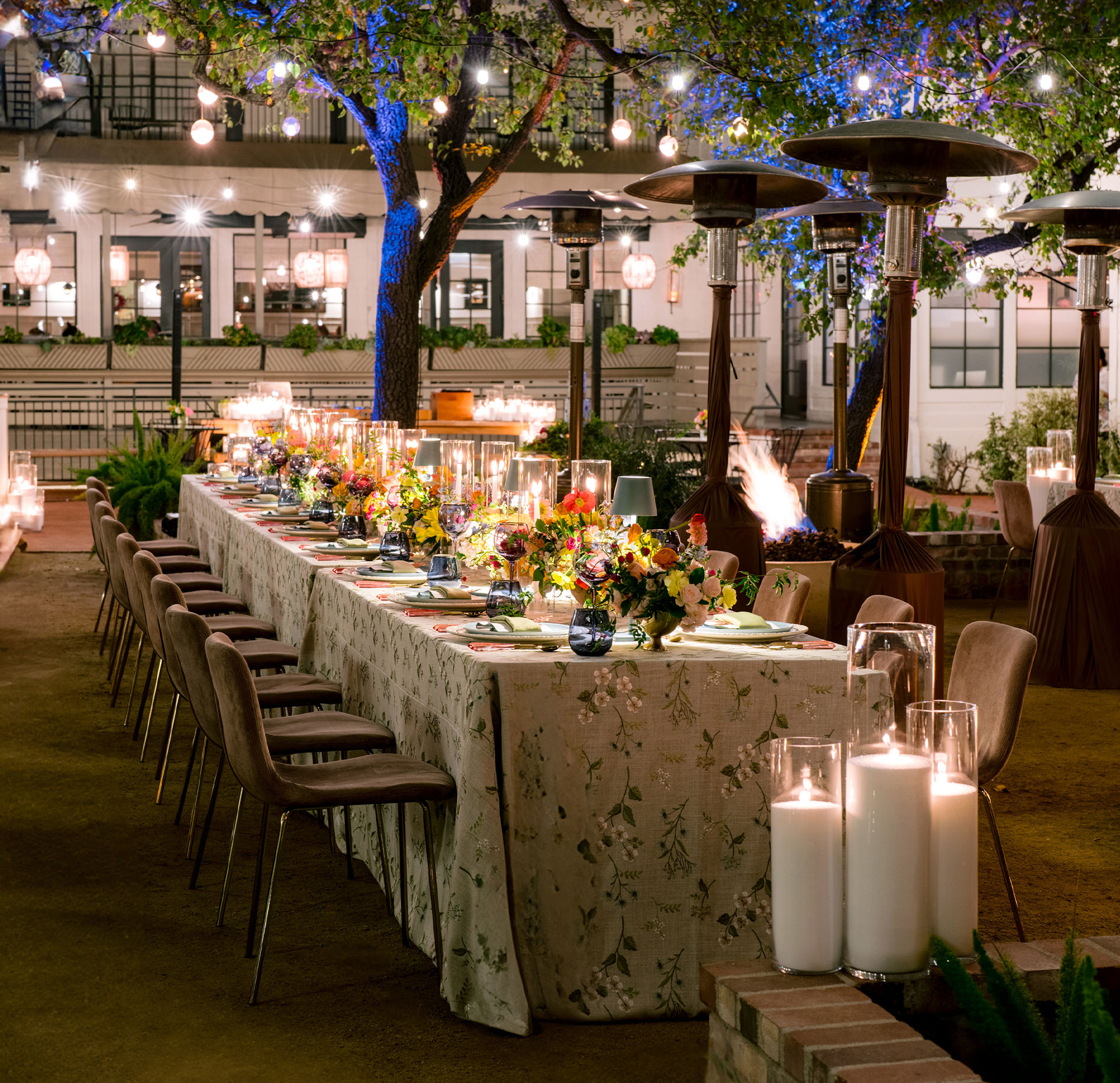 A long, elegantly set outdoor dining table with floral arrangements, candles, and cushioned chairs, surrounded by string lights and patio heaters under trees, creating a warm, festive evening atmosphere.