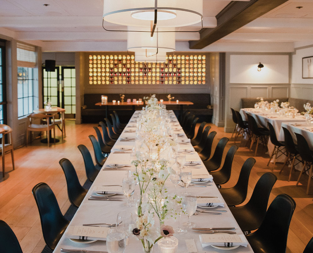 Long tables set for a formal event with white tablecloths, elegant place settings, and floral centerpieces, surrounded by black chairs in a softly lit, modern dining room—ideal for Event Spaces In Solvang.