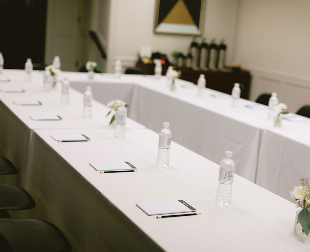 A conference room at Event Spaces in Solvang features white-clothed tables in a U-shape, each set with a notepad, pen, and bottled water. Small flower arrangements add charm to the softly lit and organized space.