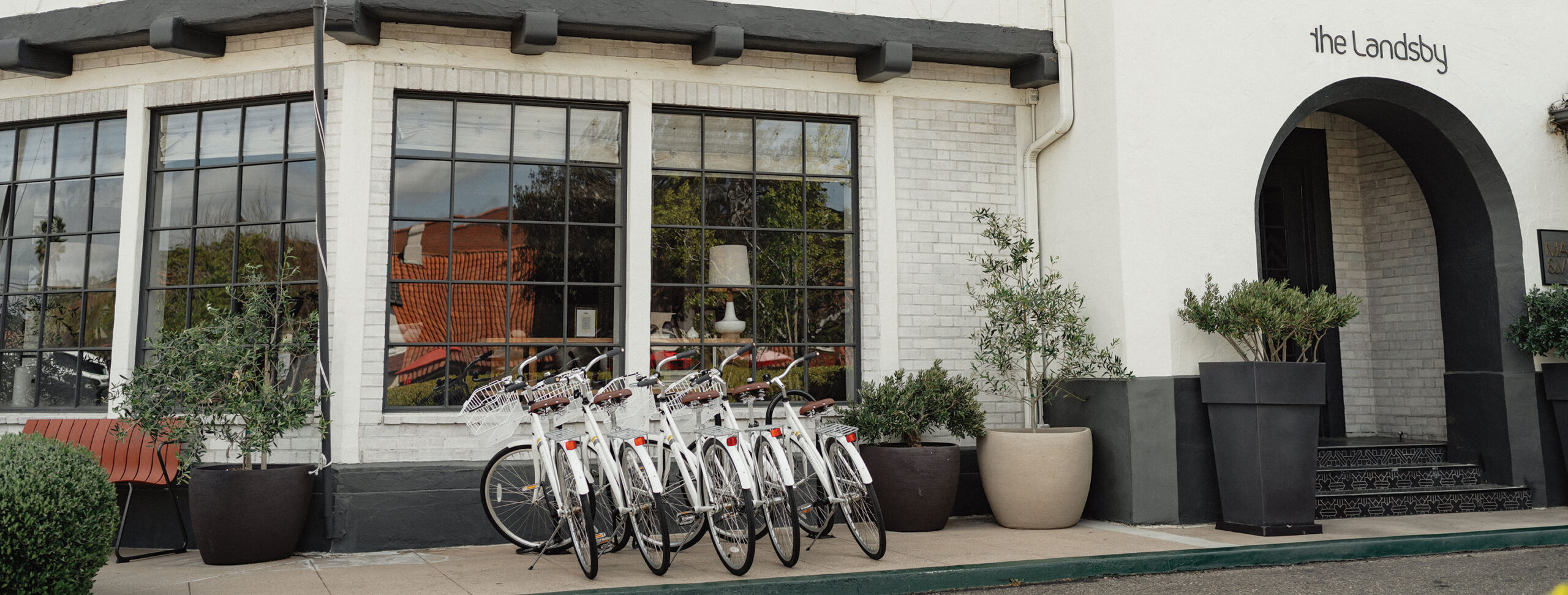 Complimentary Bikes at The Landsby A row of white bicycles is parked on the sidewalk outside a white brick building with large windows and potted plants. Discover Special Offers at The Landsby, visible above the dark archway entrance.