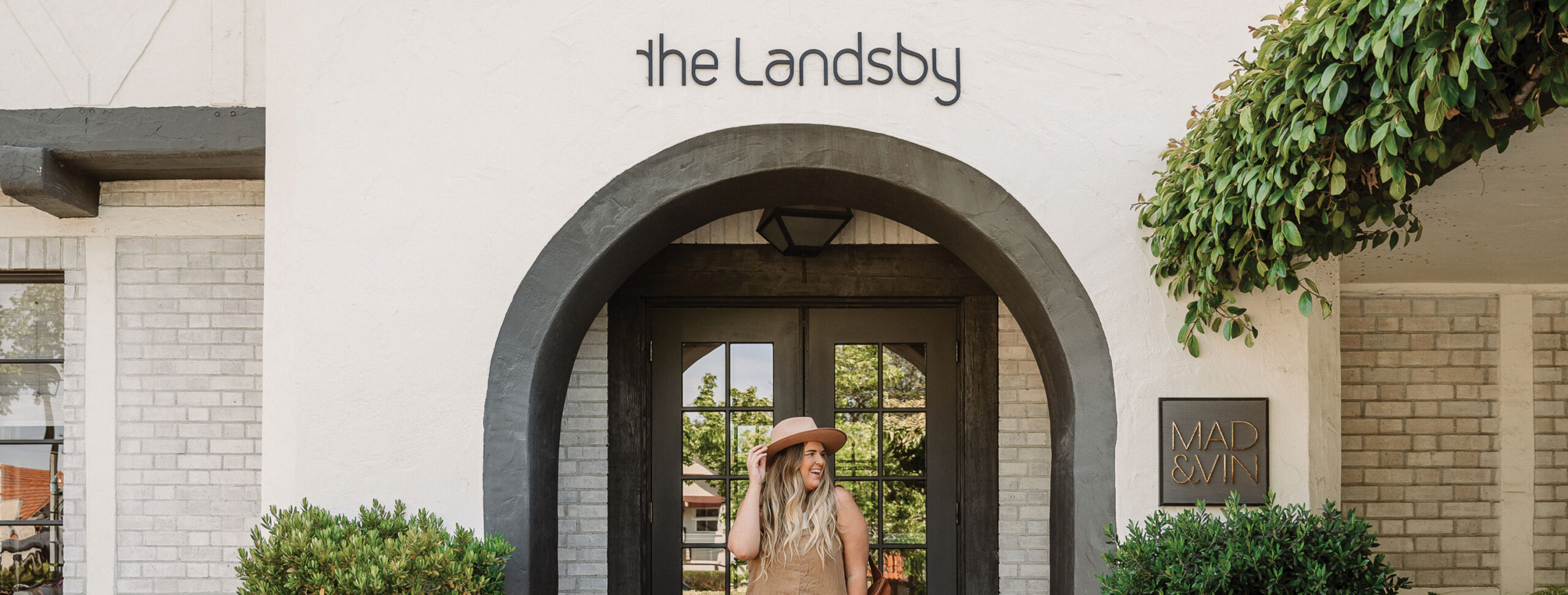 Best Hotels in Santa Barbara County A woman wearing a hat stands smiling in front of the arched entrance to a white building with a sign above the door that reads the Landsby. Green shrubs and a tree frame the entrance, hinting at its charm featured in Press & Media stories.