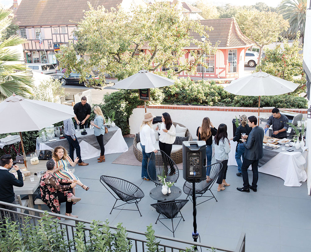A group of people gather outdoors on a patio with tables, white umbrellas, and greenery—one of the inviting event spaces in Solvang. Some stand and chat while others sit, enjoying food and drinks as sunlight filters through the trees.
