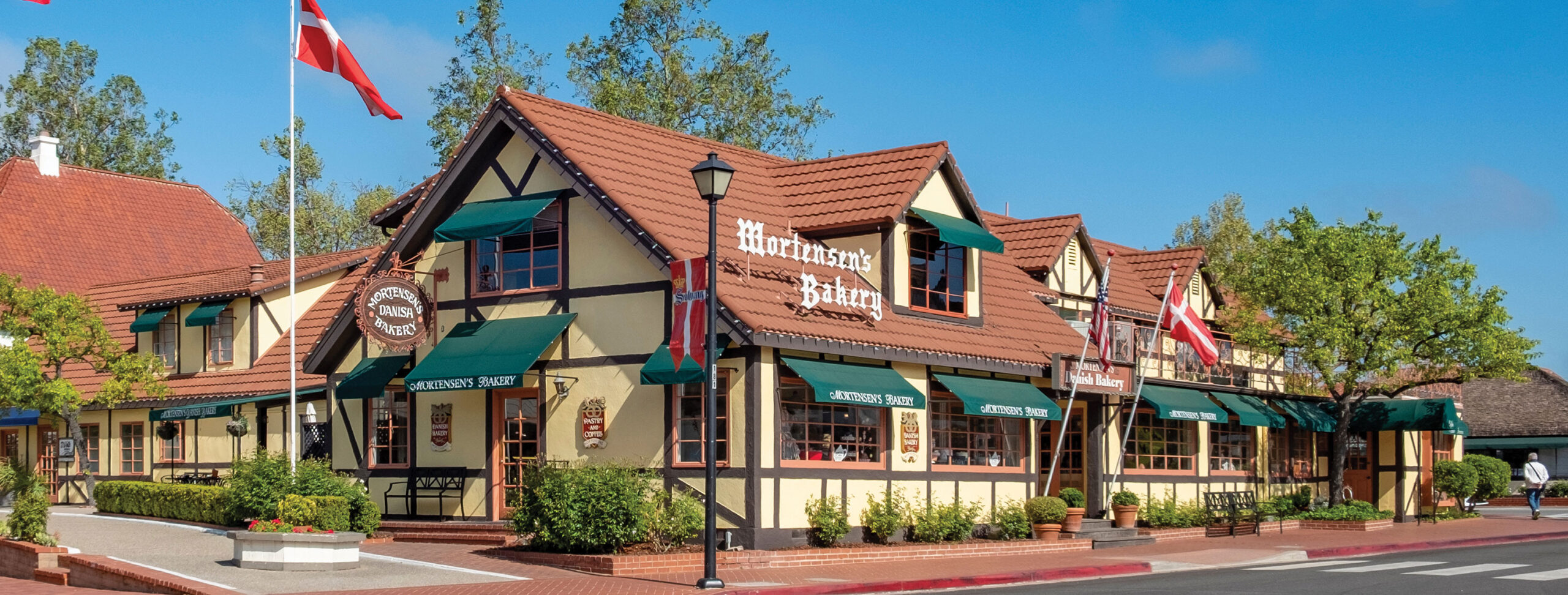 A Danish-style bakery with green awnings, half-timbered walls, and red-tiled roofs sits on a sunny street—a must-see among Things To Do In Solvang. Danish flags and signs saying Mortensens Bakery welcome visitors. Trees and blue sky surround the building.