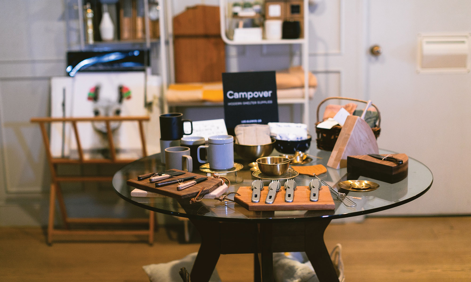 A glass table displays mugs, plates, wooden cutting boards, cutlery, and assorted kitchen items in a cozy, well-lit room at The Landsby. A sign reading Campover highlights unique Hotel Amenities at The Landsby amid shelves and paintings.