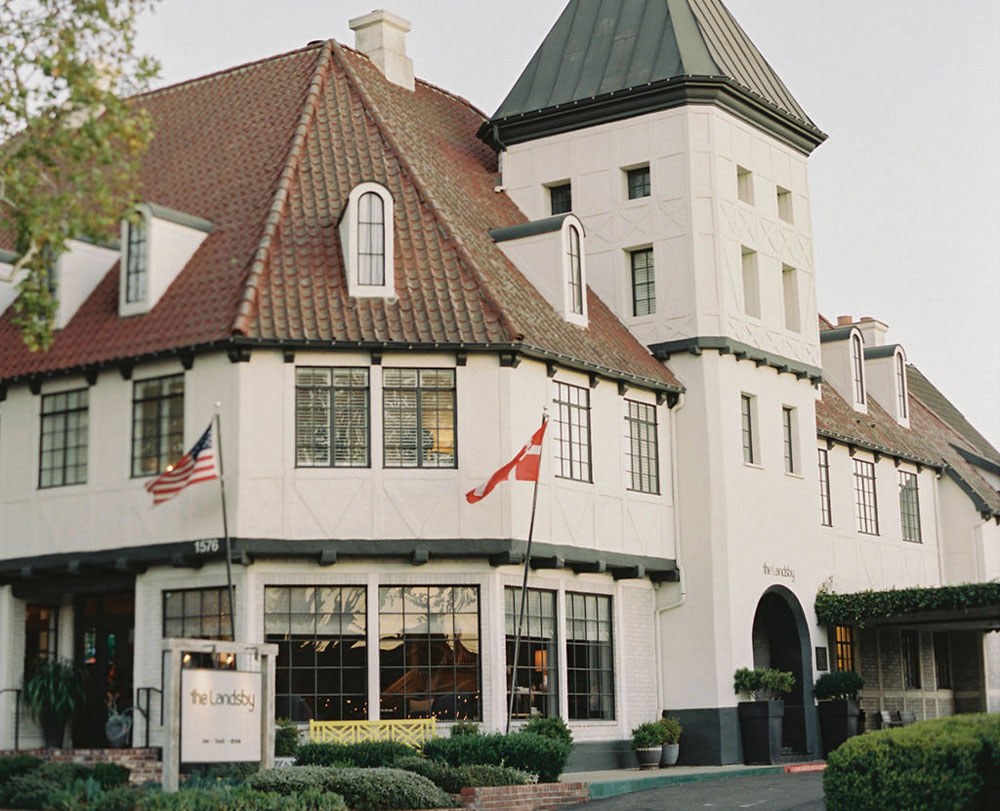 Best Hotels in Santa Ynez Valley A large, white, European-style building with a brown tiled roof, tower, and American and Danish flags by the entrance. The sign reads The Landsby. Greenery and a clear sky are visible. For inquiries, contact The Landsby.