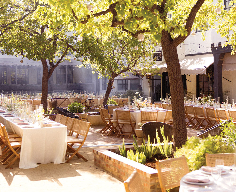 An elegant outdoor dining area in Solvang is set under leafy trees, with long tables covered in white cloths, wooden chairs, and floral centerpieces—one of the most charming event spaces in Solvang for a warm, inviting atmosphere on a sunny day.