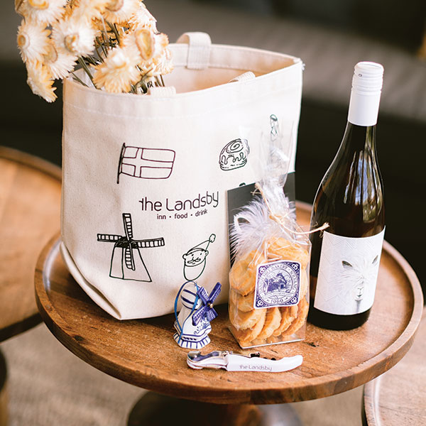 Canvas tote bag with The Landsby logo, dried flowers, a bottle of wine, cookies in a clear bag, ceramic ornament, and corkscrew, all arranged on a round wooden table—explore these delights and more with Special Offers at The Landsby.