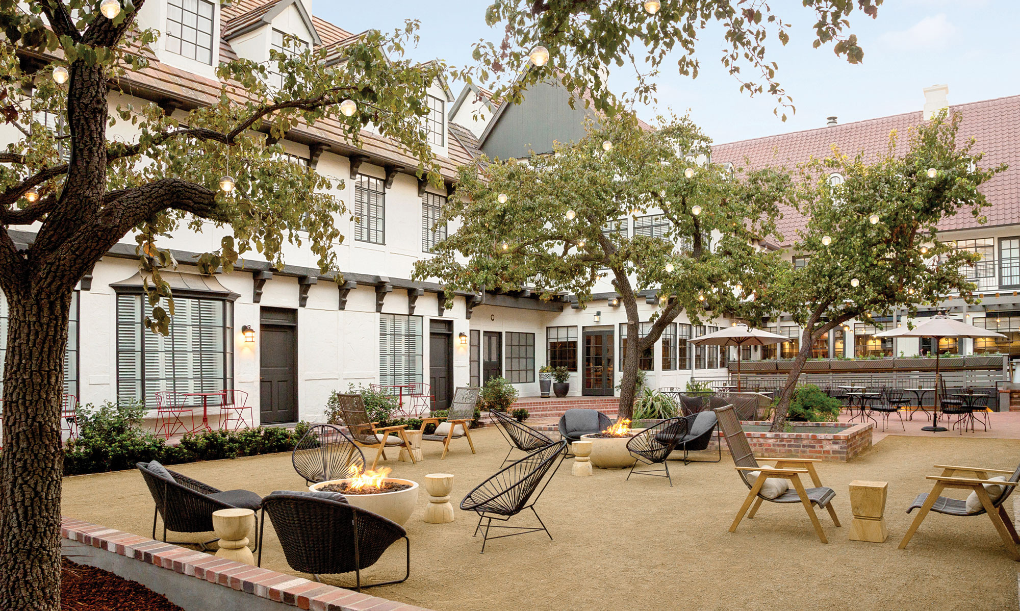 A charming outdoor courtyard showcases Hotel Amenities at The Landsby, with modern lounge chairs around fire pits, string lights in trees, and a cozy ambiance. The space is framed by buildings with white walls and red-tiled roofs.