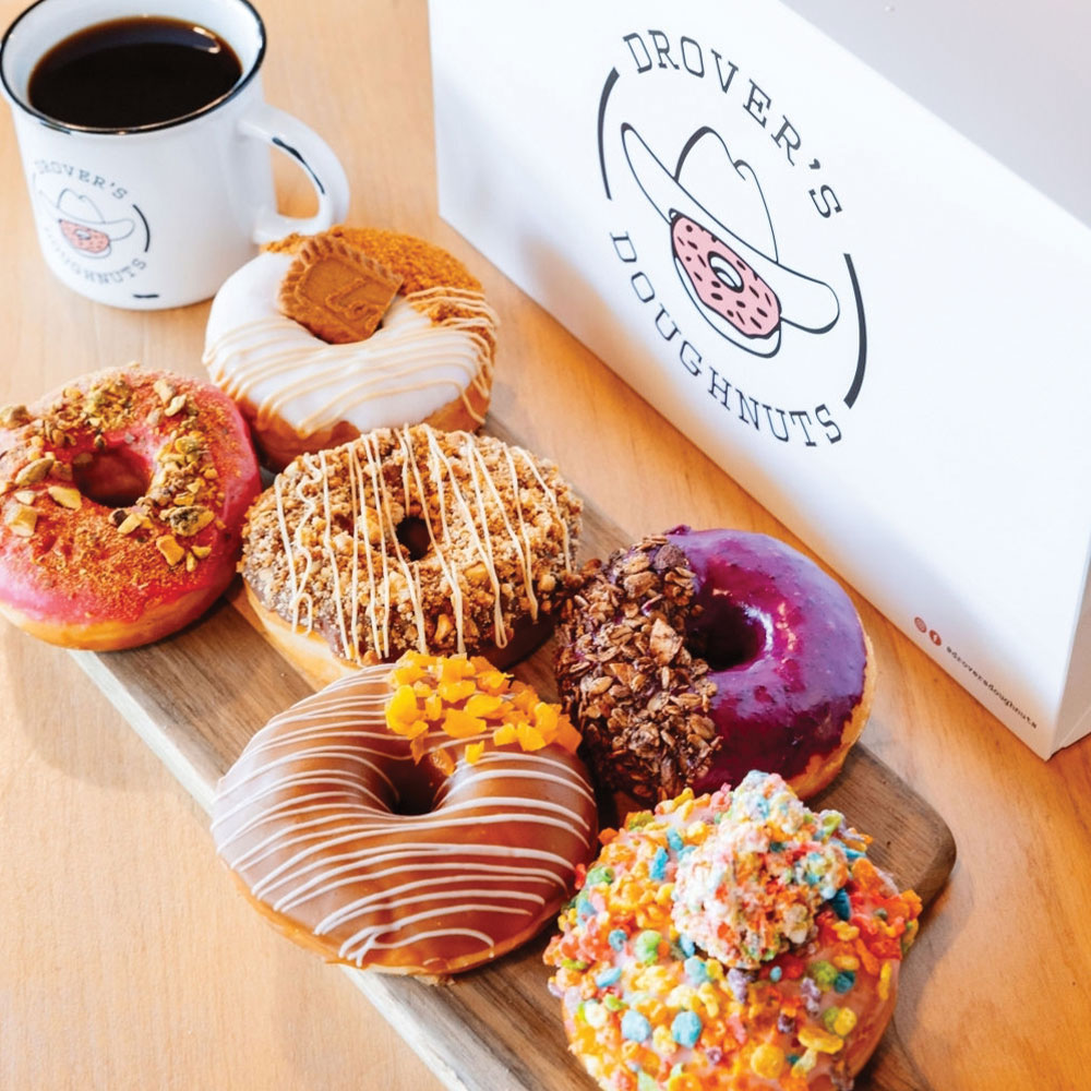 A wooden tray with six decorated donuts sits next to a white coffee mug and a Drovers Doughnuts box, topped with colorful icing. Perfect for enjoying after exploring the Local Guide at The Landsby.