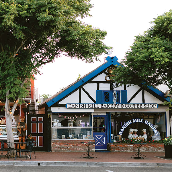 best boutique hotels in Solvang California A quaint bakery and coffee shop with a blue and white half-timbered exterior, large windows, and outdoor seating shaded by trees. The sign reads Danish Mill Bakery & Coffee Shop—a local favorite often featured in press & media.