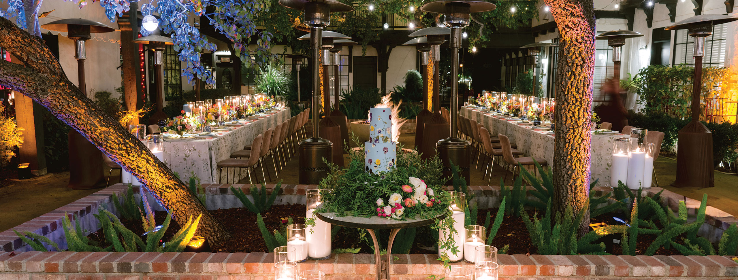 Outdoor evening event with two long banquet tables set for dinner, a decorated cake beneath trees, and string lights overhead. Candles add a warm glow to the festive atmosphere—perfect for your next Request For Proposal gathering.