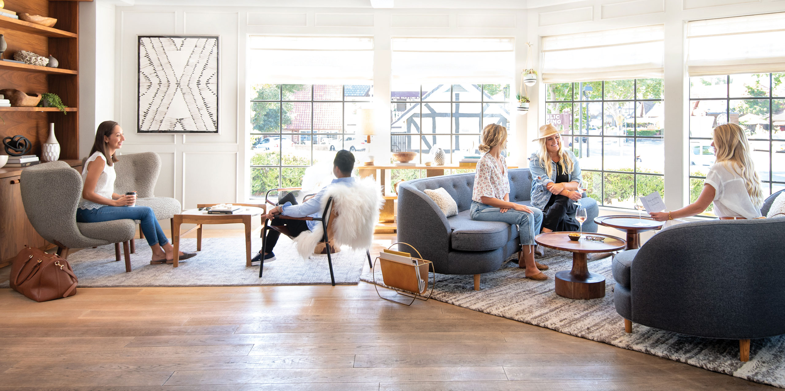 Five women sit and talk in a bright, modern living room at a boutique hotel in Solvang, with large windows, gray chairs, coffee tables, and wooden floors. Sunlight fills the space and greenery is visible outside.