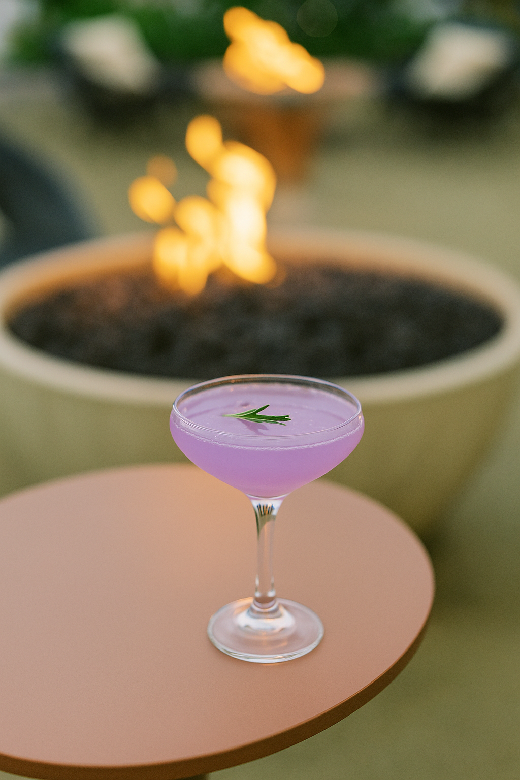 A lavender-colored cocktail garnished with a sprig of rosemary sits on a round, peach-colored table, with a large fire pit burning in the blurred background.