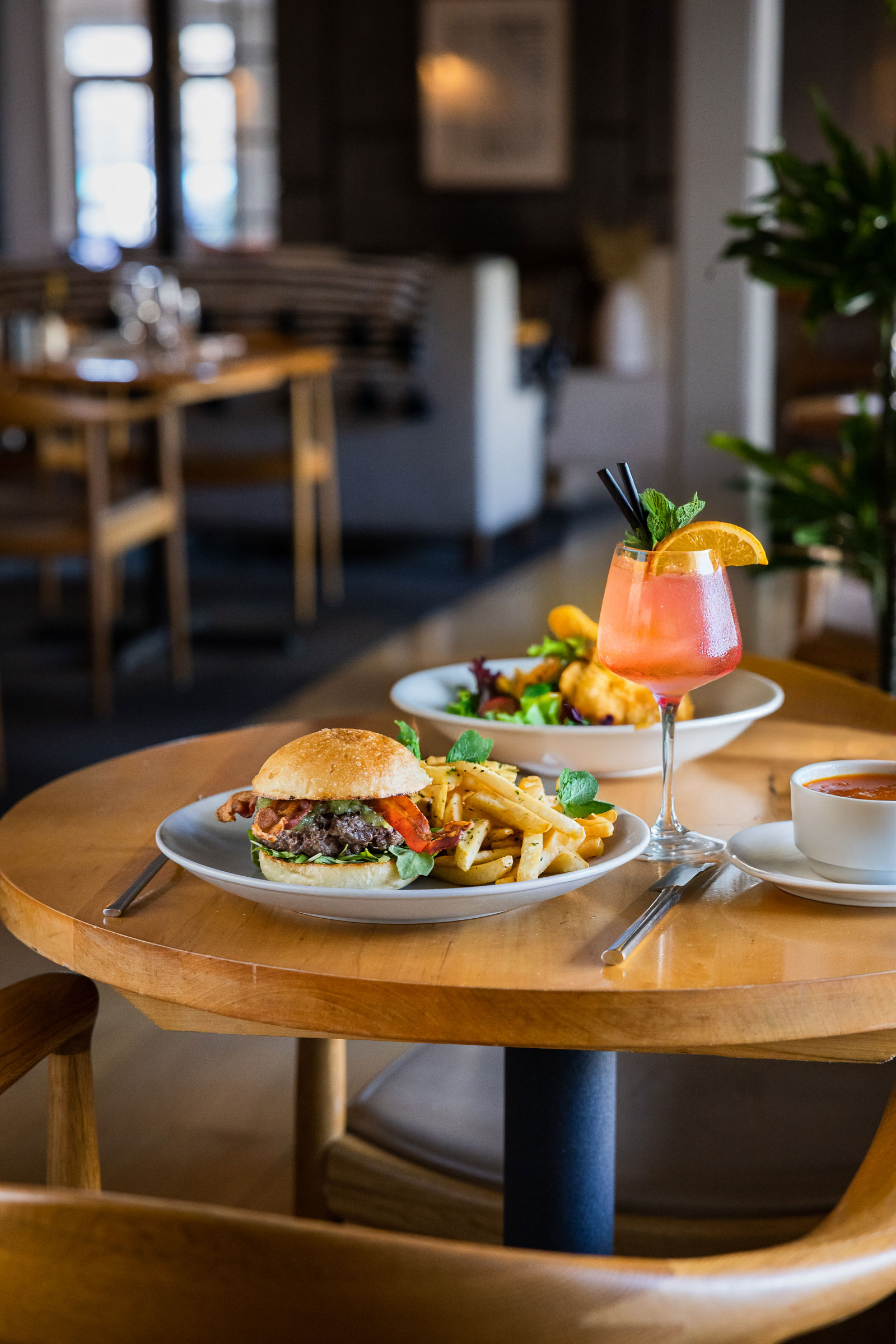 A wooden table set with a burger, fries, salad, a bowl of soup, and a pink cocktail garnished with an orange slice and mint. In the background, there are empty chairs and tables in a cozy restaurant.