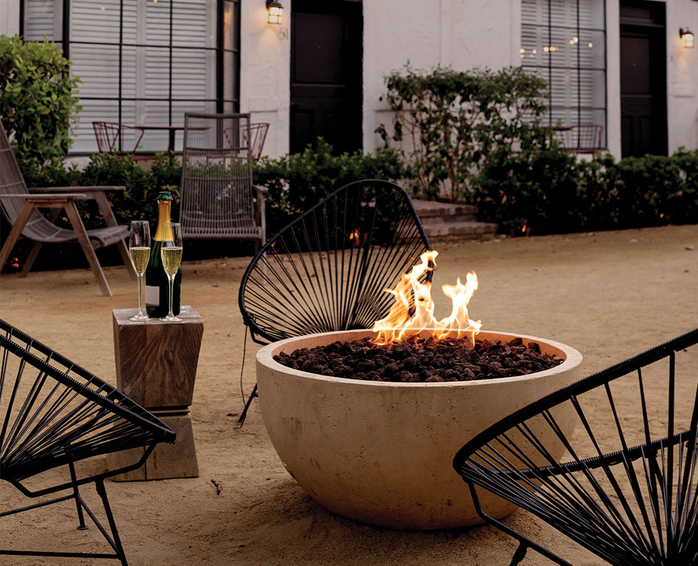 A modern outdoor fire pit with bright flames is surrounded by black metal chairs at The Landsby, a boutique hotel in Solvang. A small wooden table holds a bottle of champagne and two glasses, with bushes and a building featuring large windows in the background.