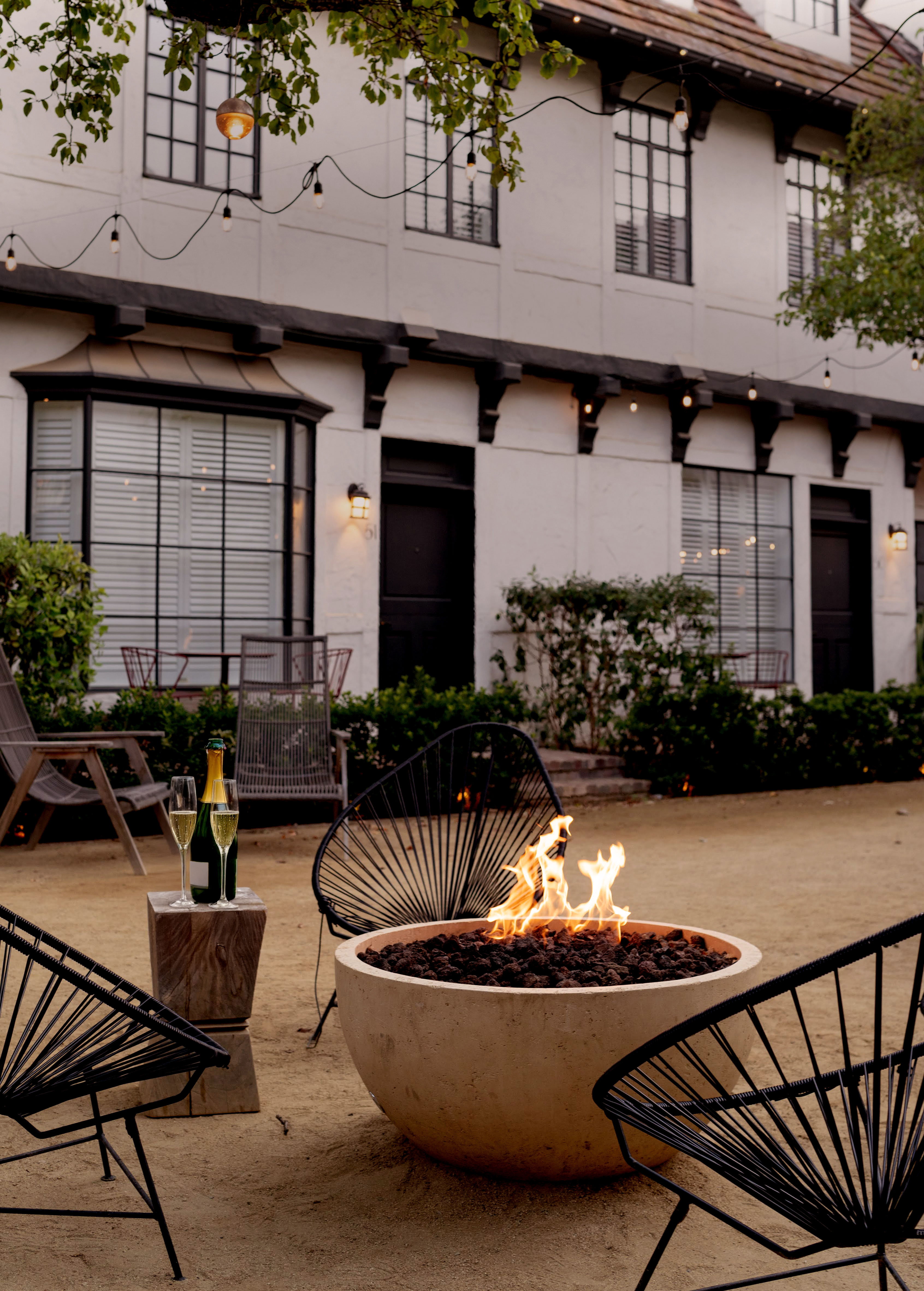A cozy outdoor patio features a round fire pit with flames, black chairs, a small table with a champagne bottle and glasses, string lights overhead, and a two-story building in the background.