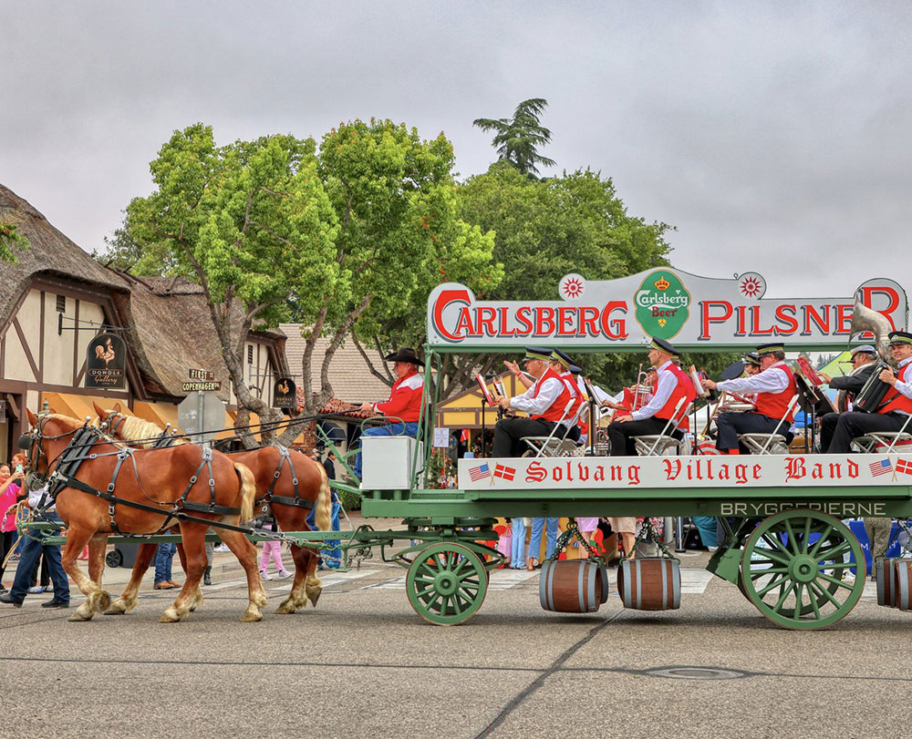 A horse-drawn wagon labeled Carlsberg Pilsner and Solvang Village Band carries musicians in red jackets, playing instruments during a parade—one of the lively Things To Do In Solvang as people and trees line the festive street during Danish Days.