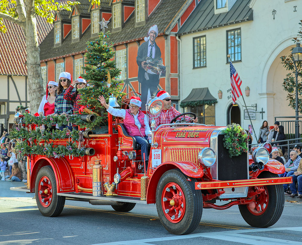 A festive red antique fire truck decorated with garlands for Julefest in Solvang, a Christmas tree, and people in Santa hats drives in a holiday parade—one of the must-see Things To Do In Solvang. Spectators watch from the street as an American flag waves in the background.