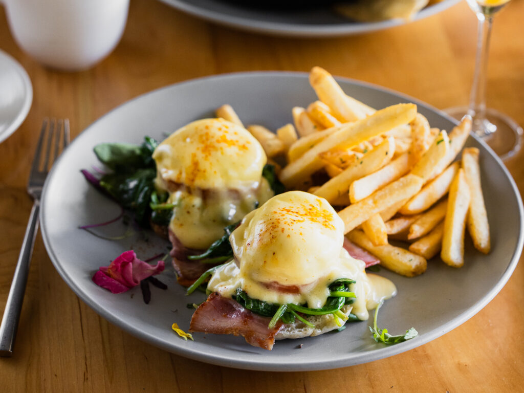 Mad & Vin page_0000_Mad&Vin_HungryInLA_Web-7 (2) A plate of eggs Benedict topped with hollandaise sauce, served with ham or bacon, wilted spinach, and golden French fries—a perfect taste of Top Solvang Dining. A pink flower garnish adds a charming touch to the presentation.