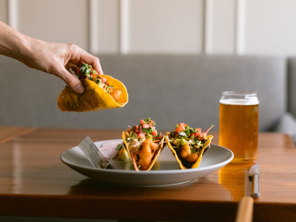 Mad & Vin page_0003_IMG_7170 (1) A hand picks up a taco from a plate with two other tacos, garnished with vegetables. The plate sits on a wooden table next to a glass of beer, showcasing Top Solvang Dining at its finest.