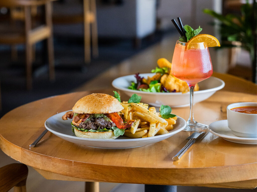 Mad & Vin page_0004_Dinner-Image A plate with a burger, fries, and greens on a wooden table, next to a glass of pink drink garnished with orange and mint, showcases Top Solvang Dining alongside bowls of soup and salad in the background.