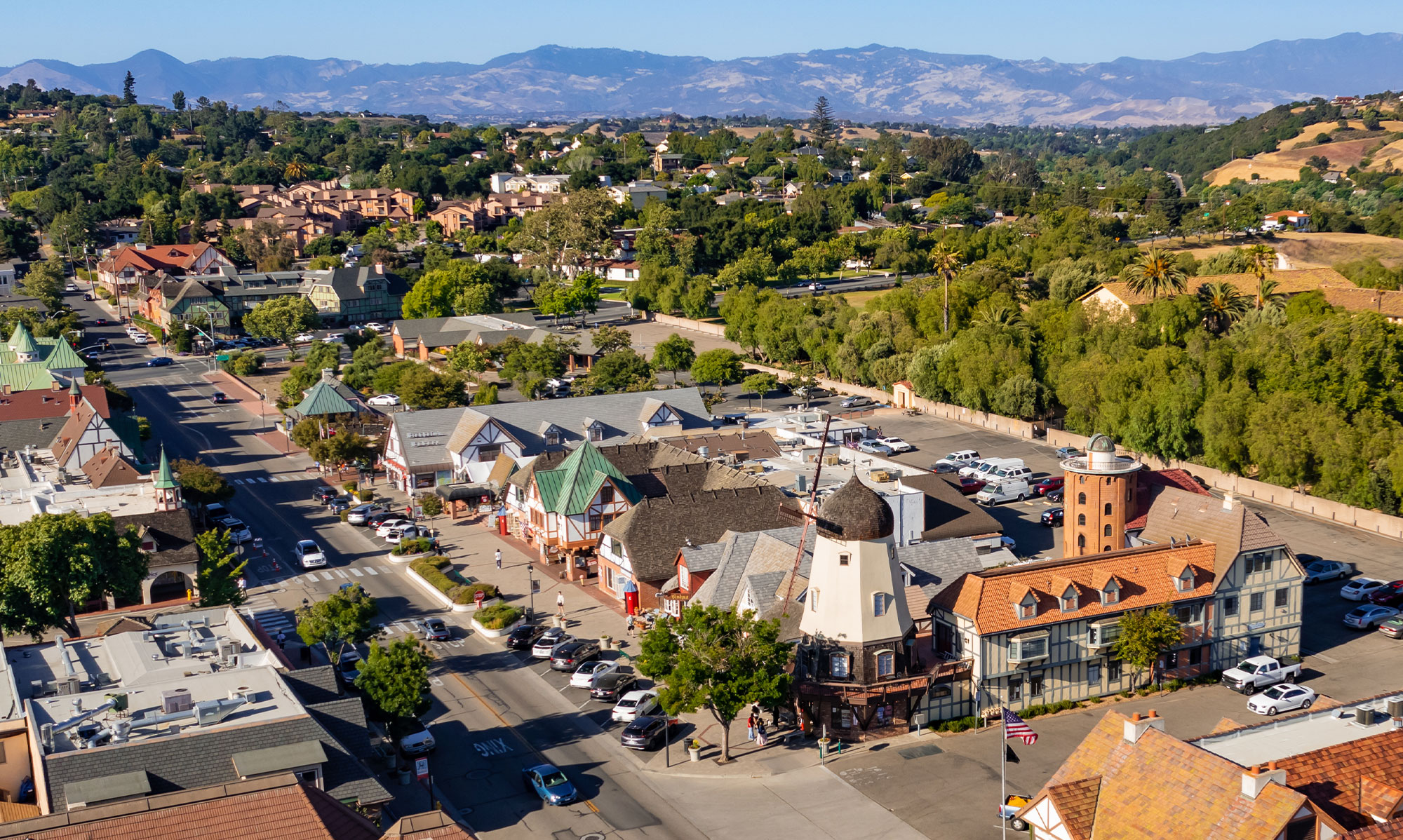 Aerial view of a small town with Danish-style architecture, featuring distinctive buildings, parked cars, tree-lined streets, and mountains in the background under a clear blue sky—perfect for exploring the many Things To Do In Solvang.