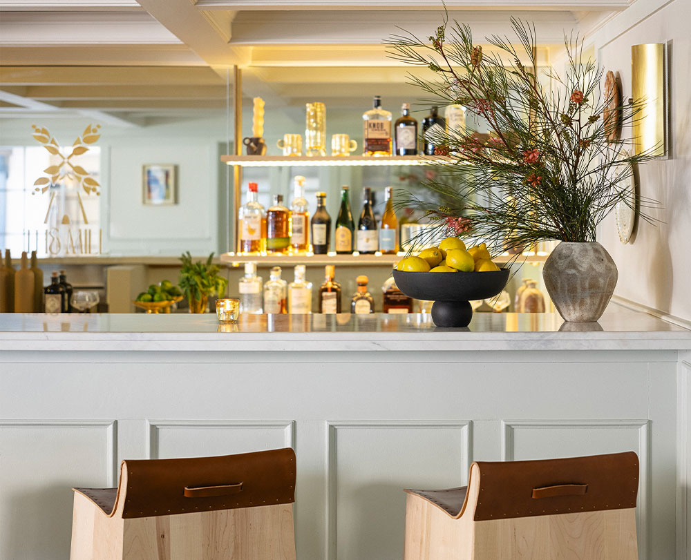 A modern bar with a white counter, wooden stools with leather backs, a black bowl of lemons, and greenery—perfect for those seeking event spaces in Solvang. Shelves in the background showcase various bottles and glassware.