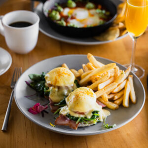dine1_0000_Mad&Vin_HungryInLA_Web-7 (2) Enjoy Top Solvang Dining with a plate of eggs Benedict featuring ham, sautéed spinach, hollandaise sauce, and fries, served alongside coffee and orange juice on a rustic wooden table.