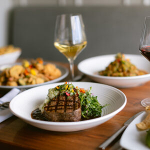 dine1_0001_IMG_7492 (2) A grilled steak served with greens and vegetables on a white plate, accompanied by glasses of wine and other plated dishes on a wooden table—perfect for experiencing Top Solvang Dining in a welcoming restaurant setting.