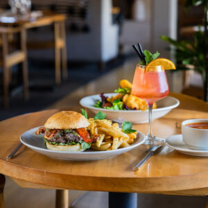 dine1_0002_Dinner-Image A wooden table set with a burger and fries, a pink cocktail garnished with orange and mint, soup, and salad in a bright, cozy restaurant—enjoy Top Solvang Dining in a welcoming setting.