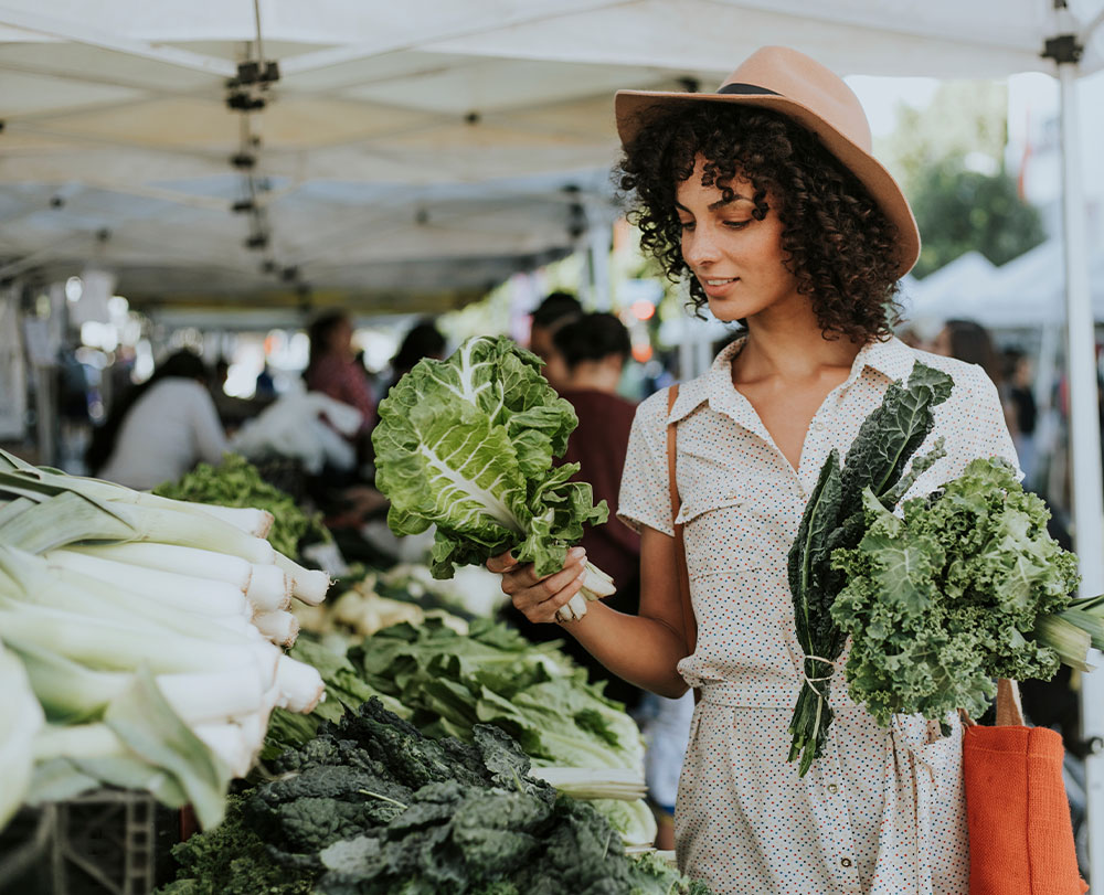 A woman wearing a hat and light dress shops at an outdoor market in Solvang, holding leafy greens in one hand and a bundle in the other—one of the delightful things to do in Solvang amid vibrant displays of fresh vegetables.