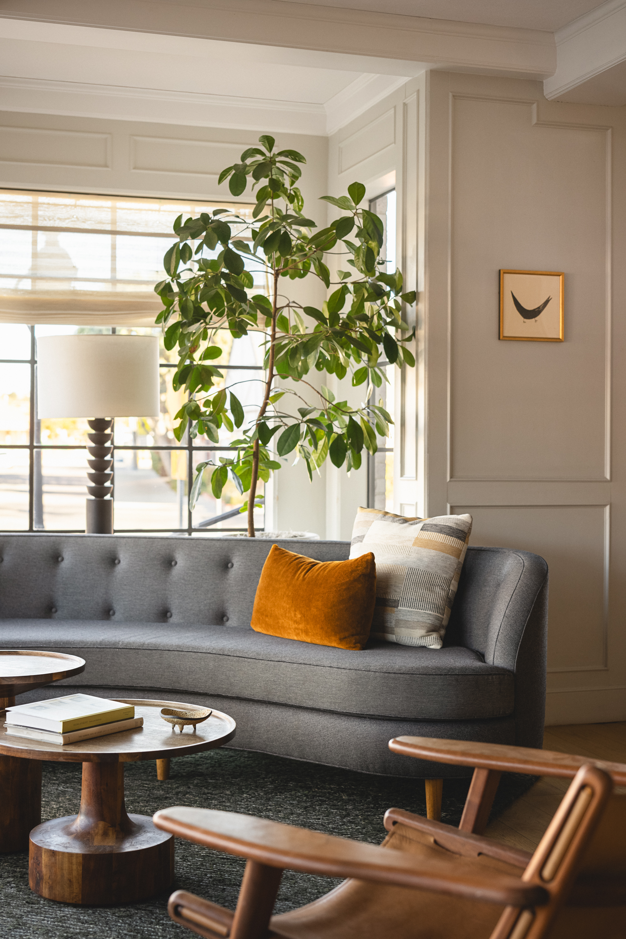 The Landsby lobby with a gray tufted sofa, orange and striped pillows, wooden coffee tables, a large leafy plant, a tall lamp, and a framed minimalist art piece on the white paneled wall. Sunlight streams through large windows.