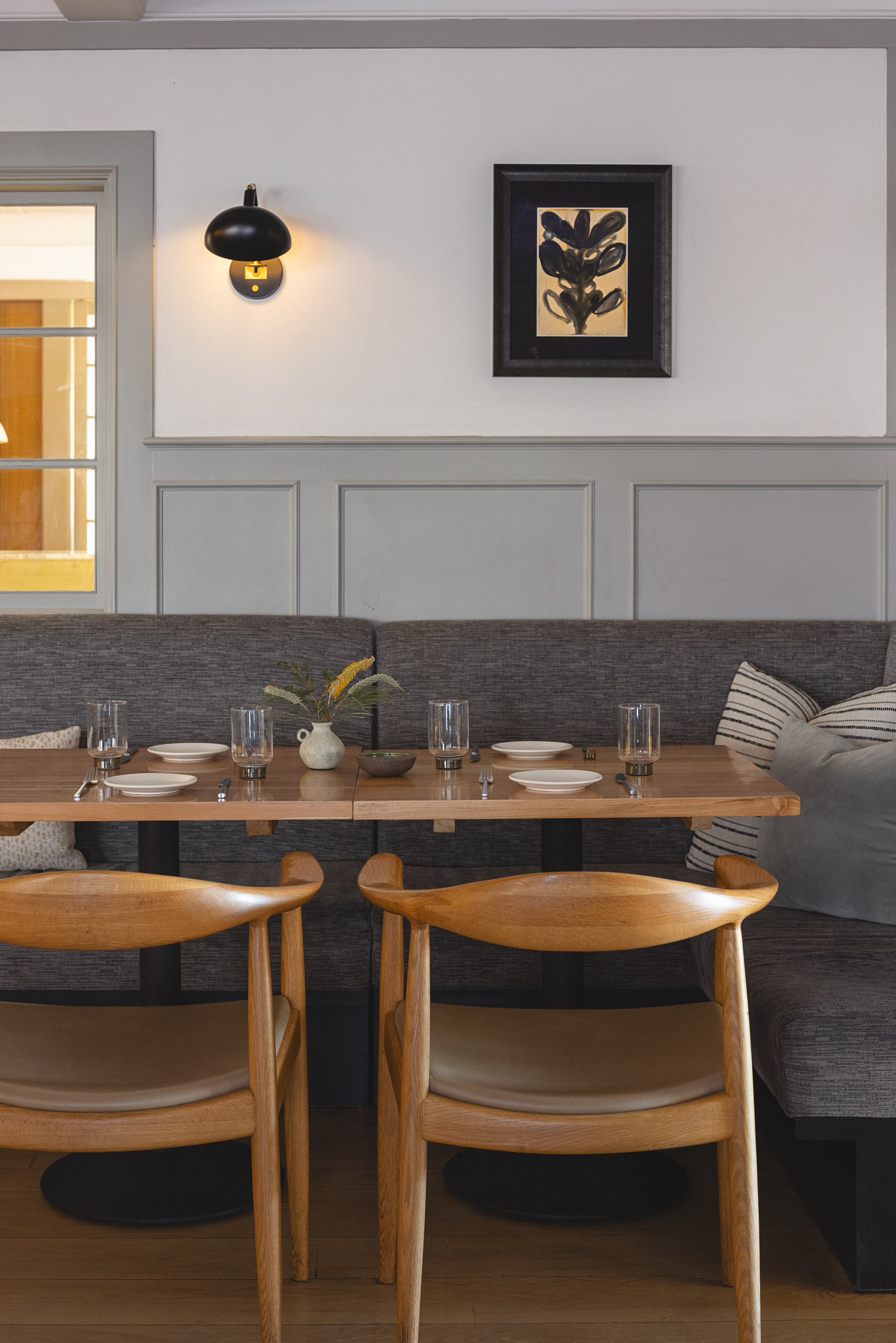 A cozy restaurant interior with two wooden chairs at a set table, gray cushioned bench seating, minimalist decor, a wall sconce, and framed art above paneled gray walls.