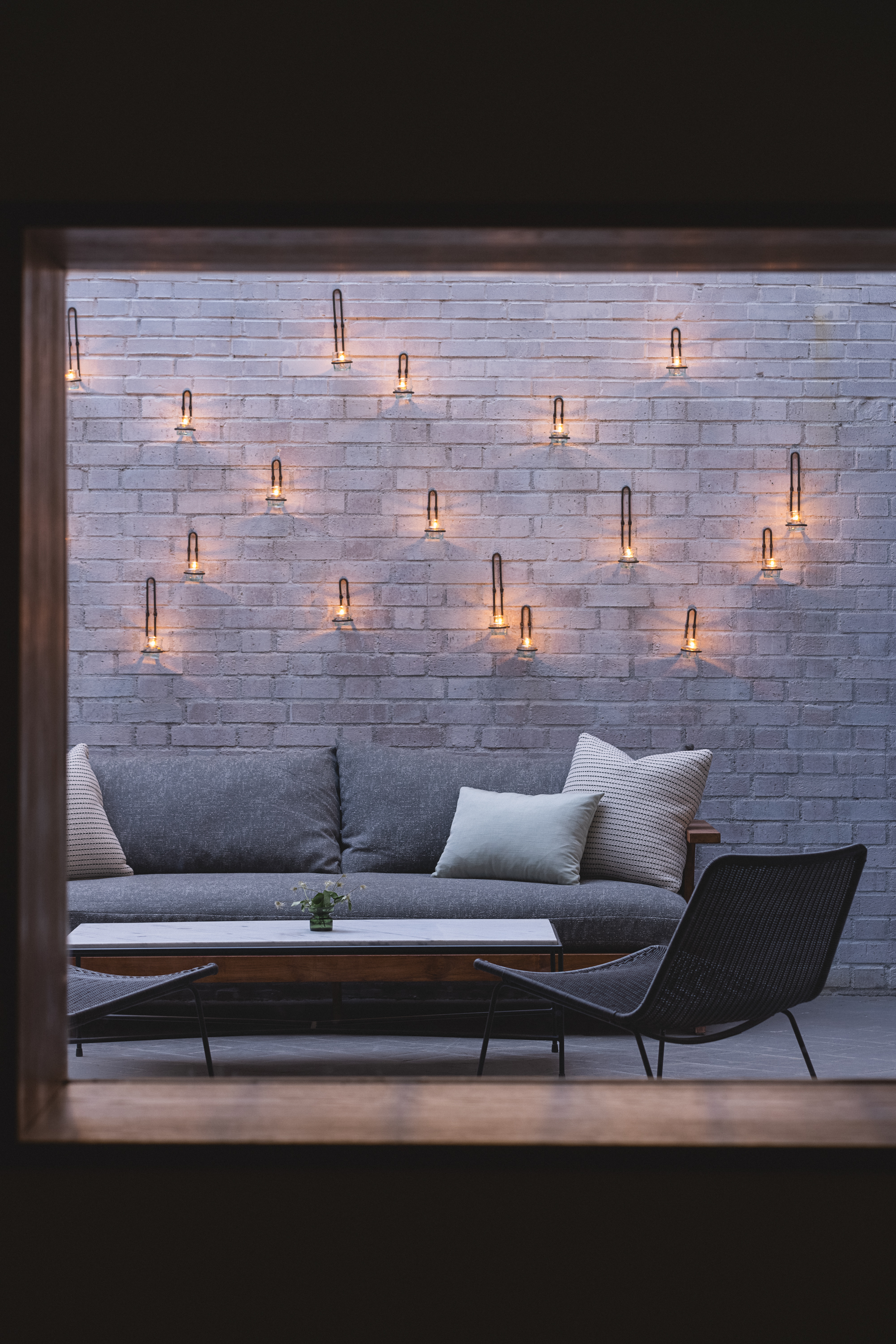 A cozy outdoor seating area with a gray sofa, accent pillows, a black chair, and a coffee table, set against a brick wall decorated with several small, glowing wall-mounted lamps.