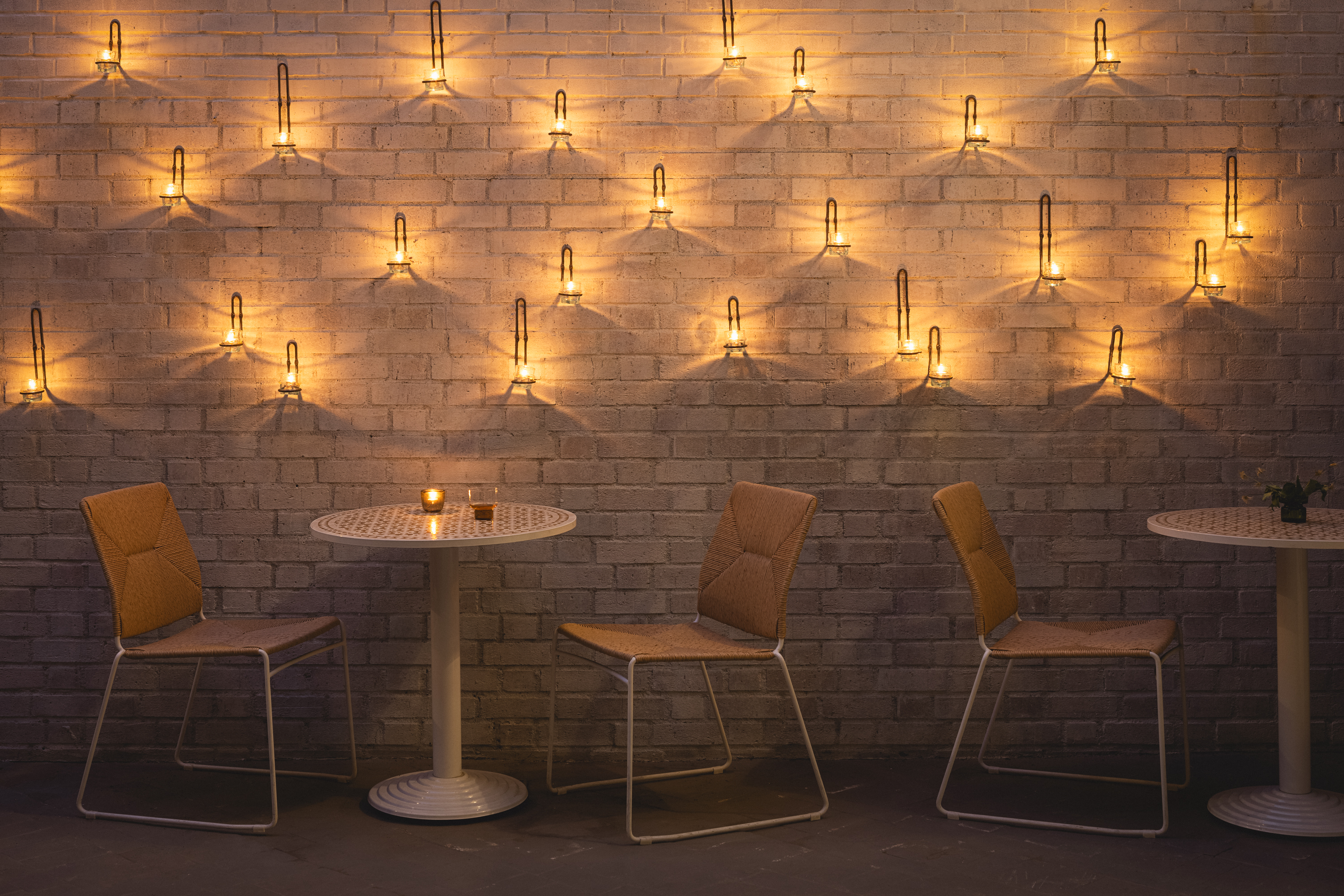 Three empty chairs and two tables are set against a brick wall adorned with rows of glowing wall lamps, creating a warm and cozy ambiance in the dimly lit café.