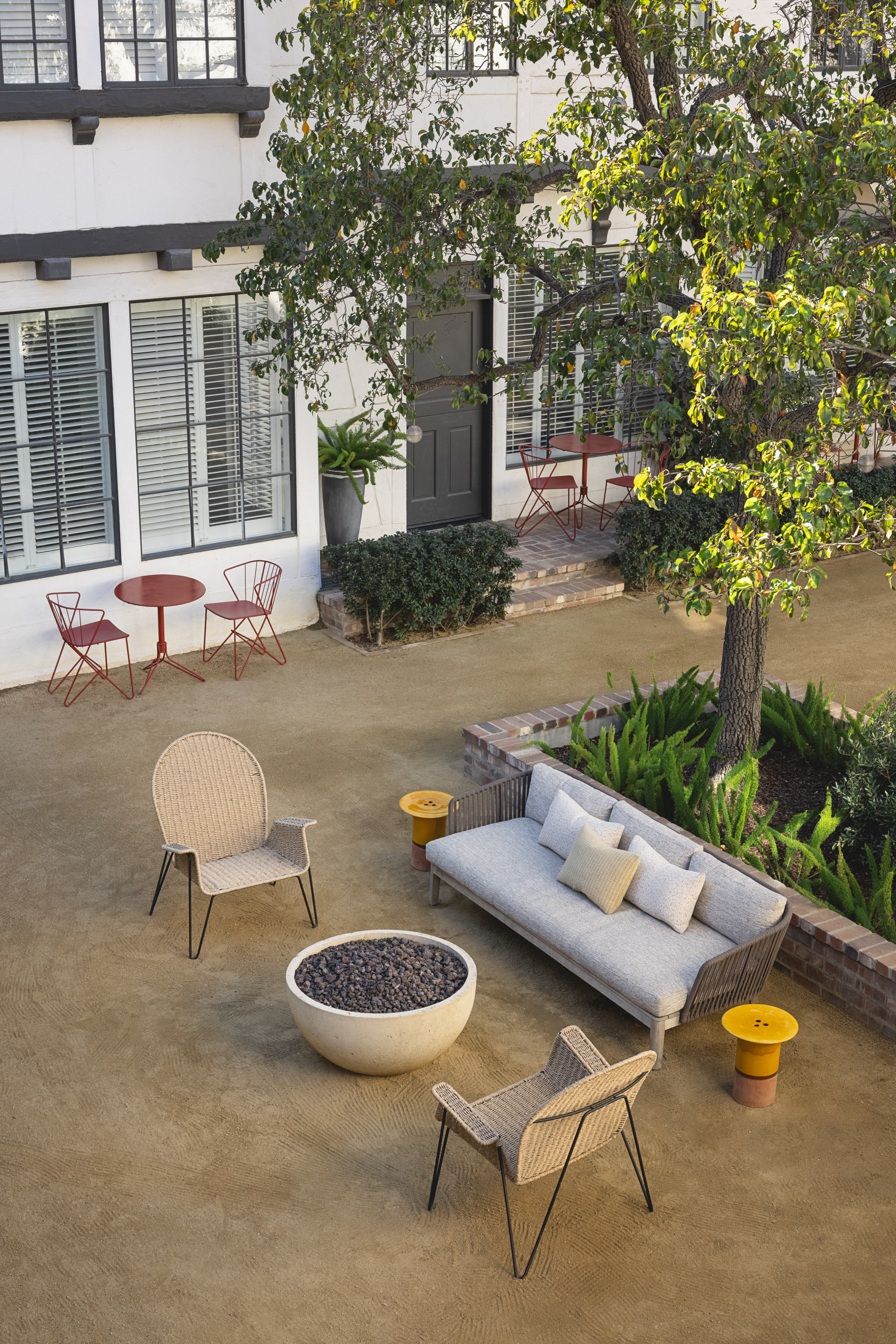 A patio with a sofa, two chairs, and a round fire pit sits under a tree. Nearby, a red bistro table with two chairs is set by a building with large windows and plants along the wall.