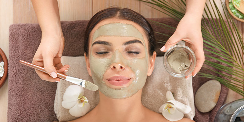 A woman with closed eyes lies on a towel as a green facial mask is applied to her face with a brush. Surrounded by orchids and spa items, she enjoys one of the relaxing things to do in Solvang.