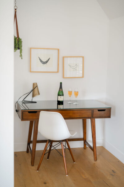 A wooden desk with a glass top holds a bottle of champagne and two filled glasses, ready for a Sparkling Celebration. A modern lamp sits on the desk, while minimalist framed artworks hang above. A white chair is positioned in front.