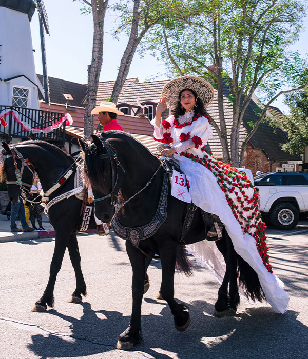 A woman in a white dress with red flowers and a large hat waves while riding a black horse in a parade—one of the festive things to do in Solvang. A man in a red shirt and cowboy hat rides another horse beside her along tree-lined streets.