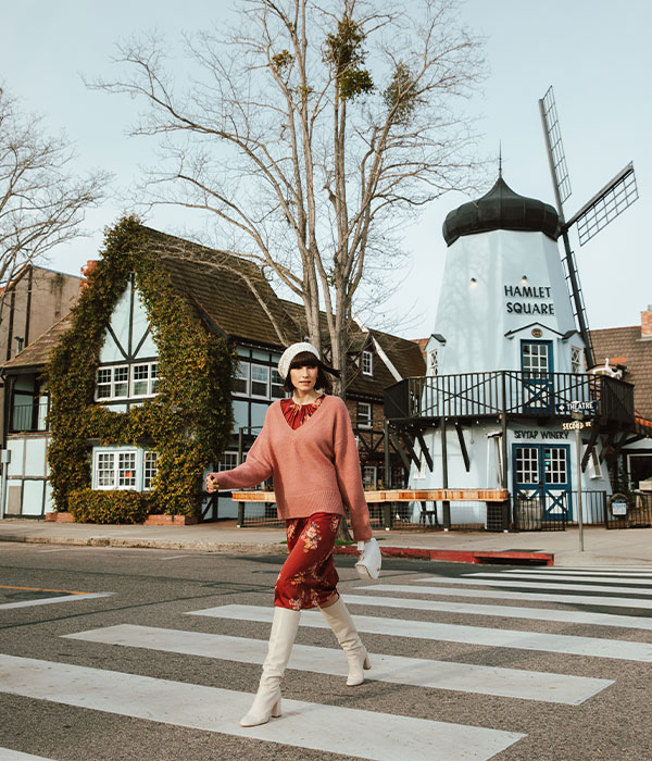 A woman in a pink sweater, floral skirt, and white boots walks across a crosswalk in front of European-style buildings, including a windmill, at Hamlet Square—one of the charming Things To Do In Solvang on a clear day.