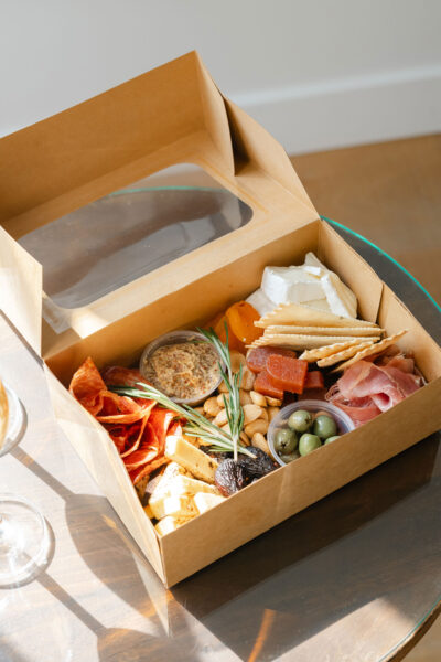 A cardboard box from Grazing Hour containing a selection of charcuterie items—crackers, cheeses, cured meats, olives, nuts, dried fruits, pâté, and fresh herbs—sits atop a round glass table.